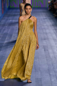 Model walks the runway in a shimmering gold one-shoulder gown with black strap, paired with strappy sandals.