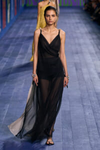 Female model walking a fashion runway in a sheer black wrap dress with thin straps and sandals, jewelry on both wrists.