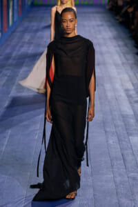 Model walking a fashion runway in a floor-length, semi-sheer black gown with gathered shoulders and long ribbon-like sleeves; red accents peek from the sleeves.
