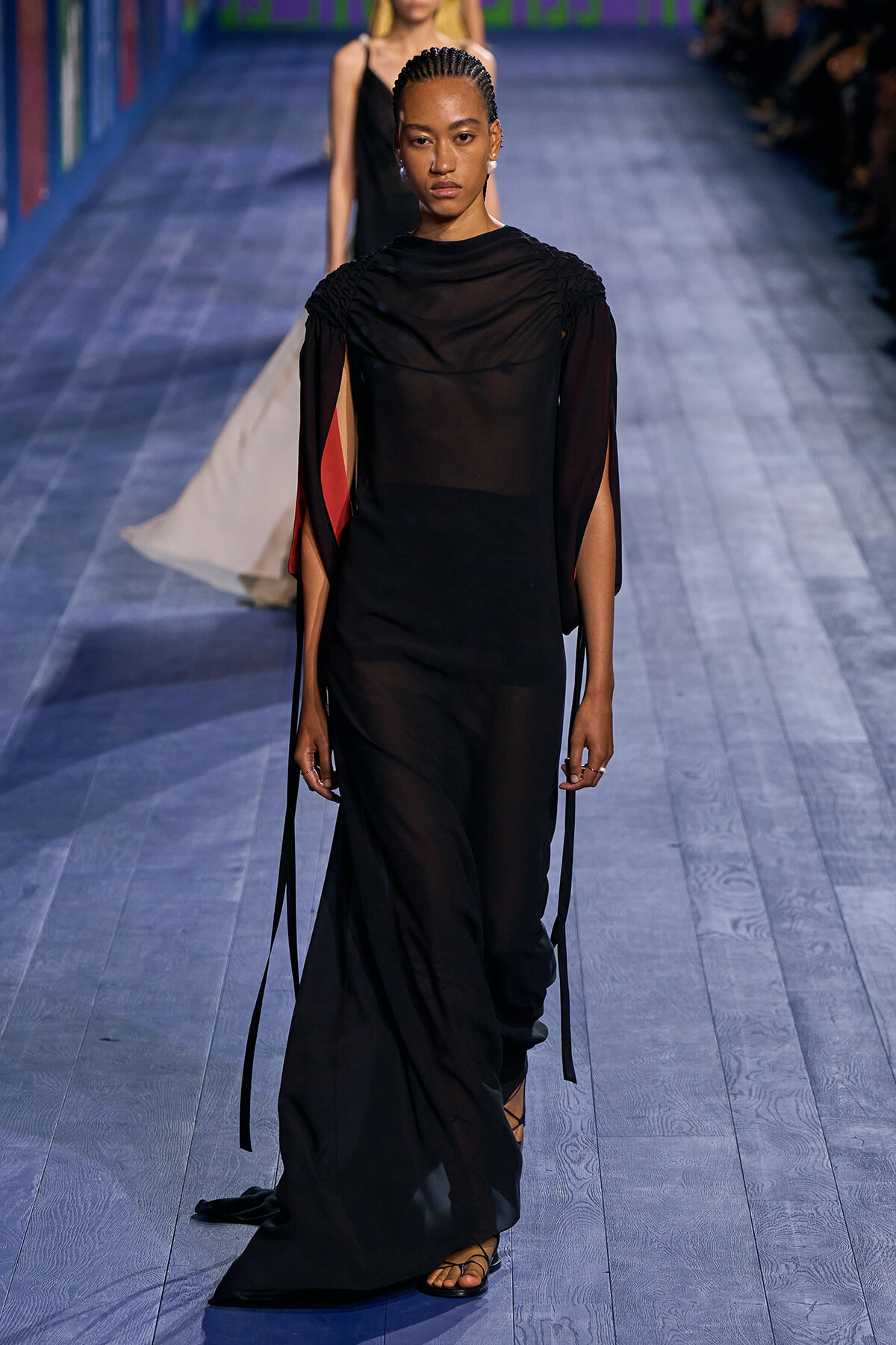 Model walking a fashion runway in a floor-length, semi-sheer black gown with gathered shoulders and long ribbon-like sleeves; red accents peek from the sleeves.