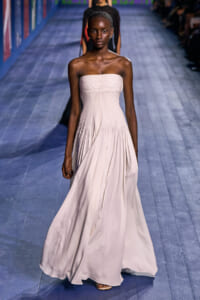 Model walks a pale pink, strapless, floor-length gown down a blue wooden runway at a fashion show.