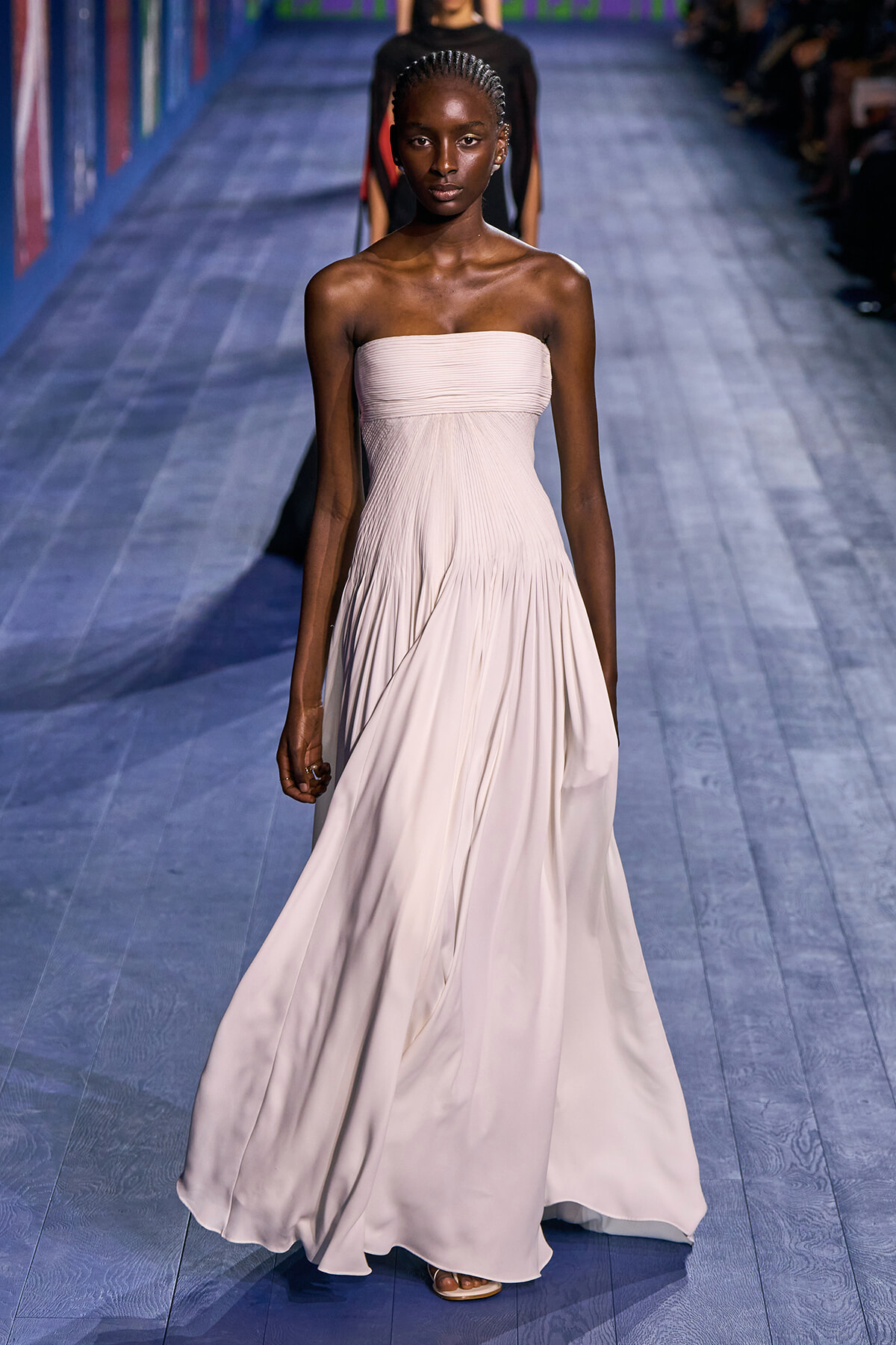 Model walks a pale pink, strapless, floor-length gown down a blue wooden runway at a fashion show.