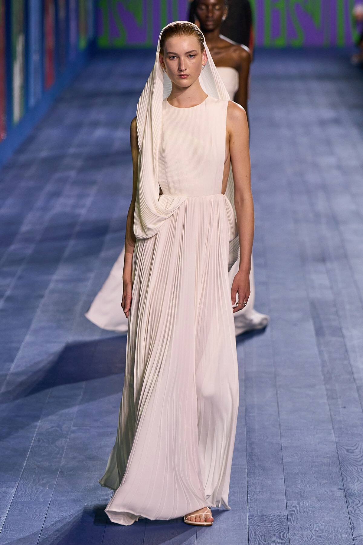 Model walking runway in a flowing ivory pleated gown with a matching veil draped over her head.