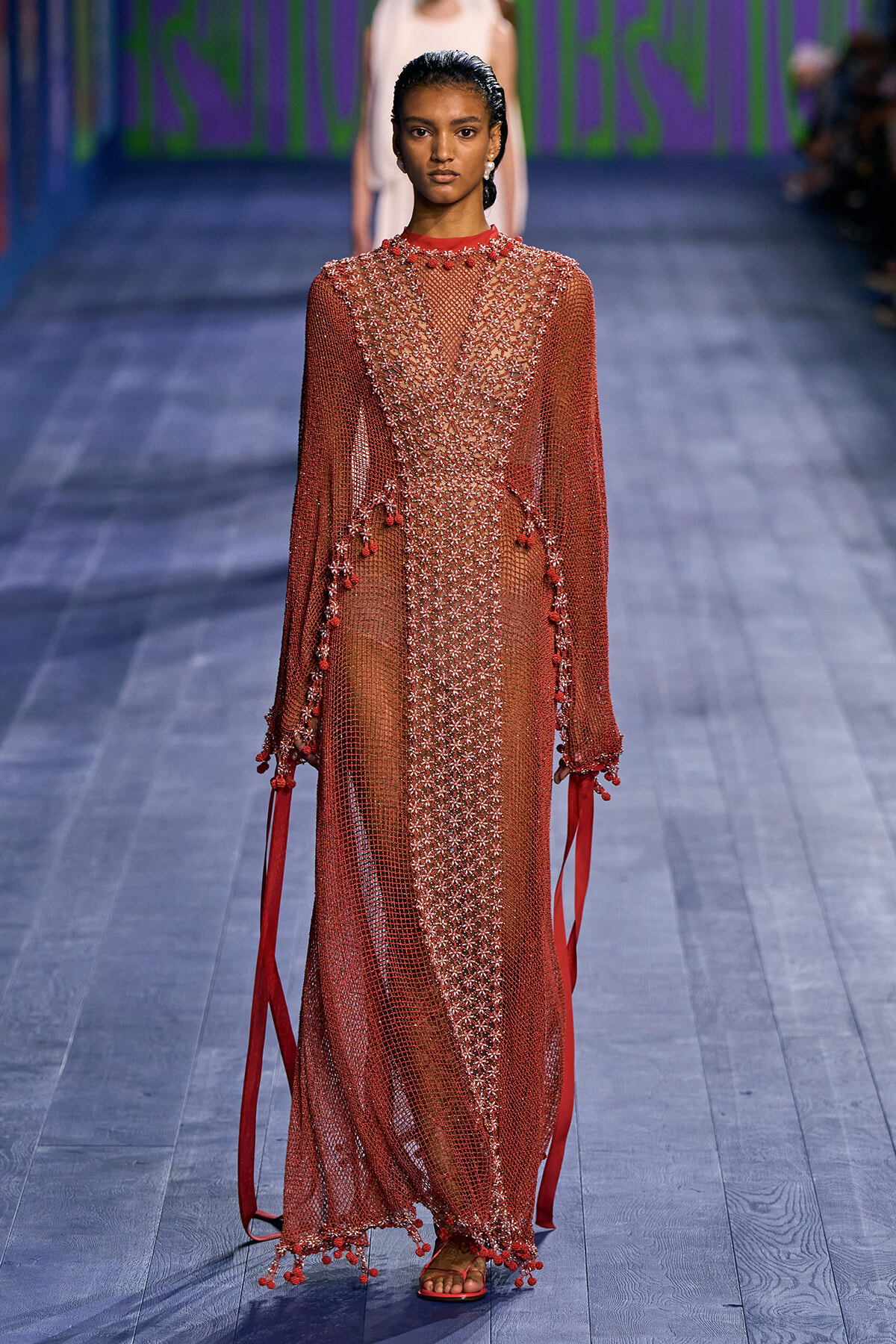 Model walking the runway in a full-length sheer red gown with intricate bead embroidery and long sleeves, red fringe details at cuffs and hem, and strappy sandals.