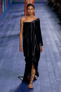 Runway model in a one-shoulder black velvet gown with a draped front and thigh-high slit, wearing lace-up sandals.