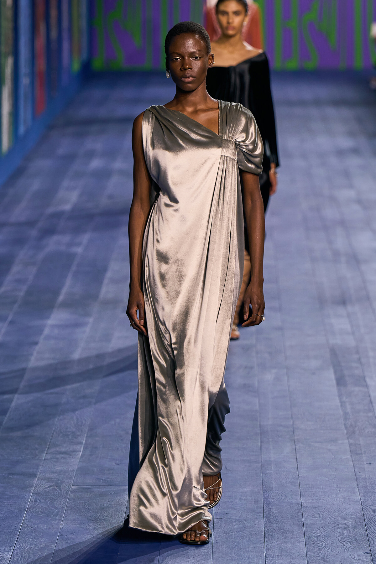 Model walking a fashion runway in a silver satin, one-shoulder draped gown with gathered detail at the chest.