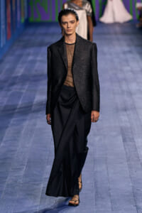 Model walks the runway in a glittery black blazer over a sheer brown top and draped black trousers with strappy sandals.