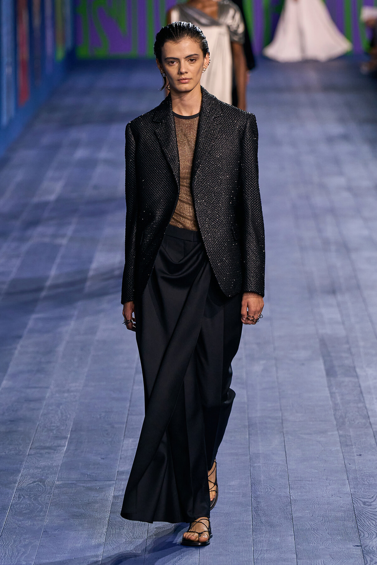 Model walks the runway in a glittery black blazer over a sheer brown top and draped black trousers with strappy sandals.