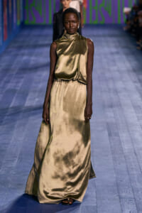 Model walks the fashion runway in a floor-length gold satin dress with a high neckline and cinched waist.