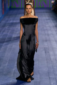 Model walking a runway in a black off-the-shoulder satin gown with minimal jewelry and strappy sandals, blue-toned floor beneath.