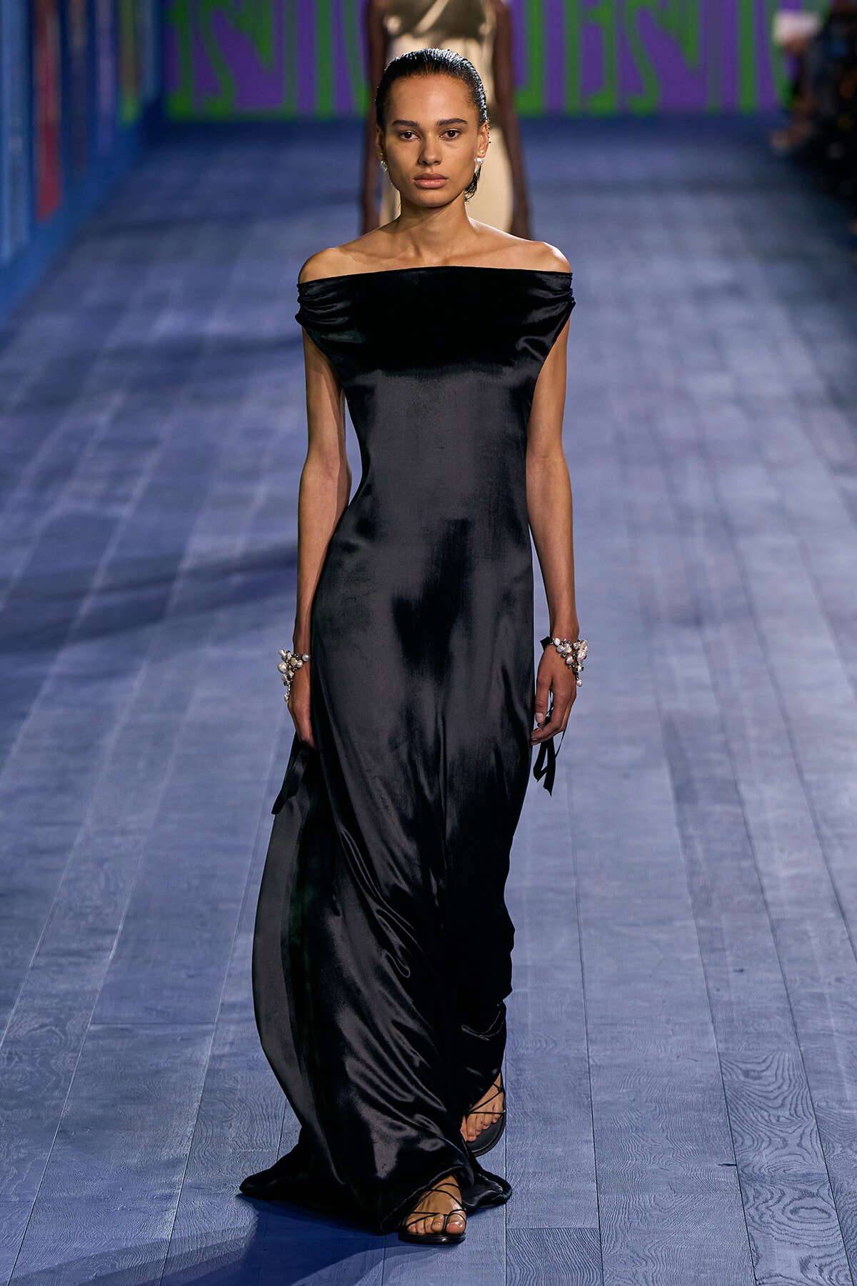 Model walking a runway in a black off-the-shoulder satin gown with minimal jewelry and strappy sandals, blue-toned floor beneath.