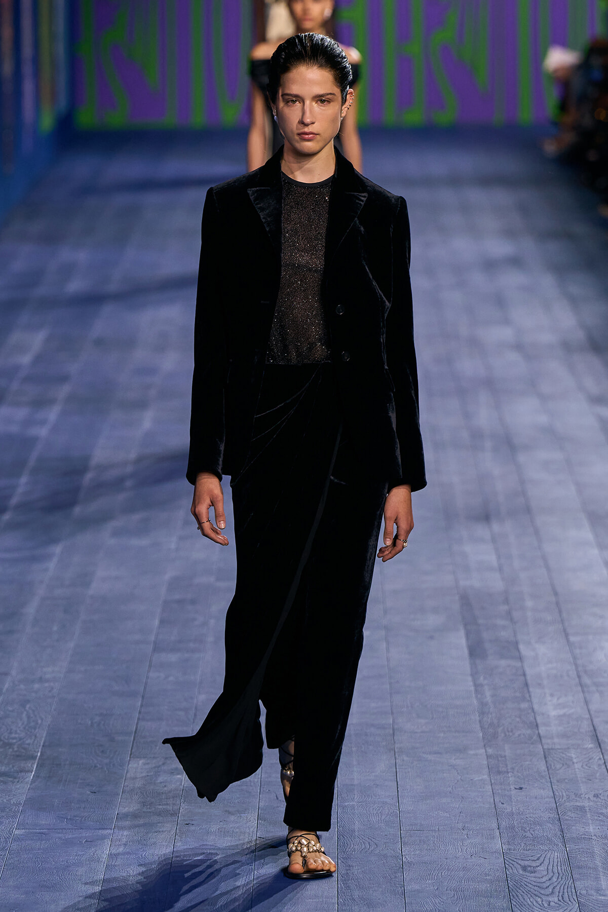 Model walks a runway in a black velvet blazer and matching pants with a sheer shimmer top under the blazer.