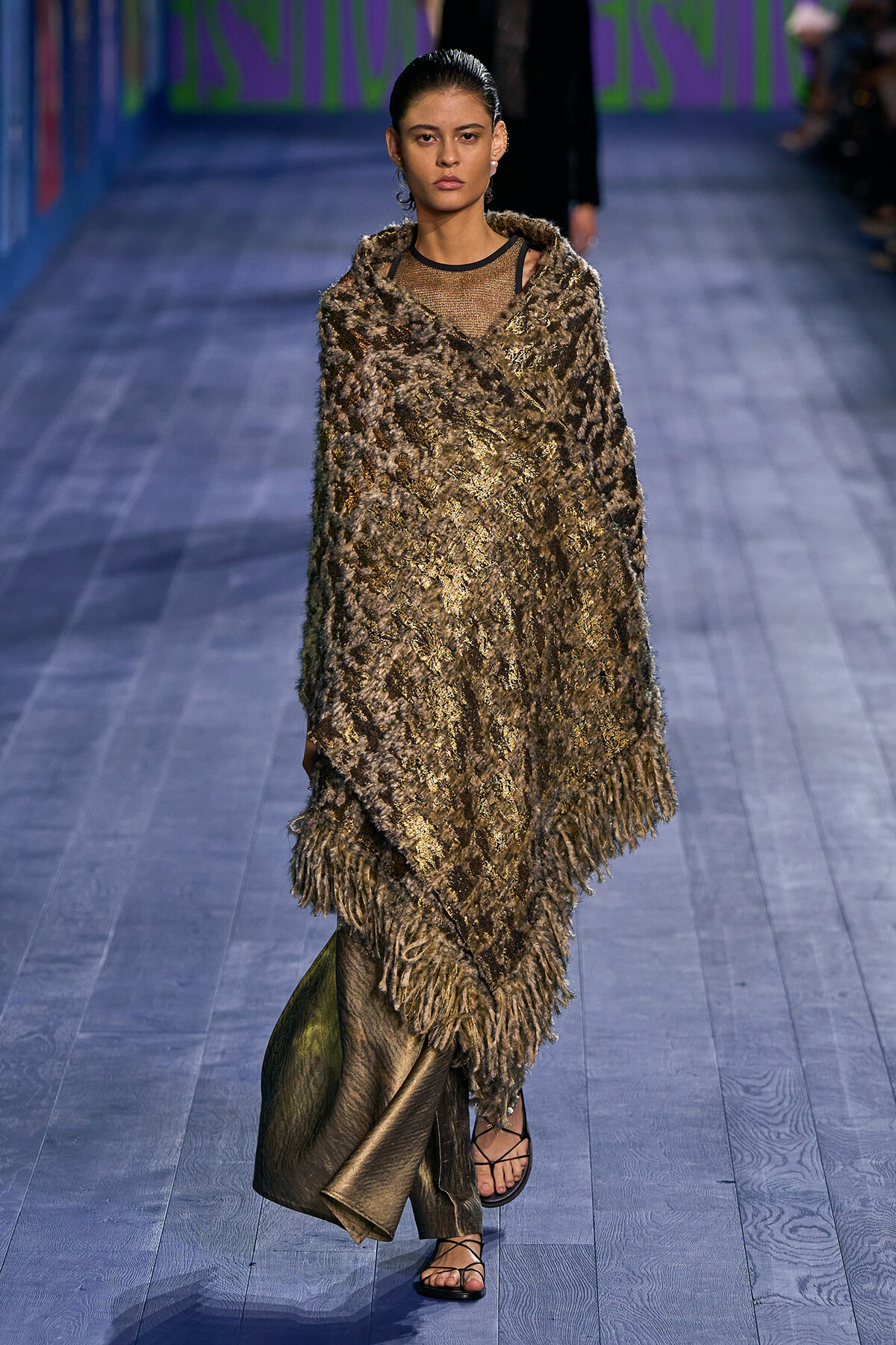 Model walks down runway wearing a large textured brown fur wrap over a sheer top, paired with a metallic skirt and strappy sandals.
