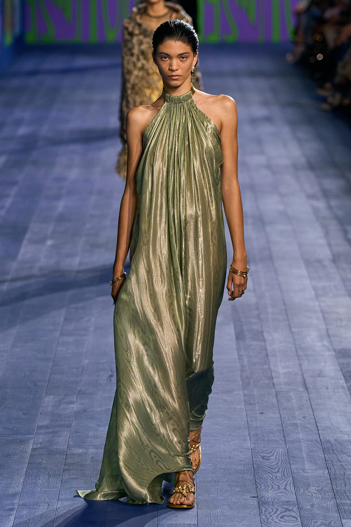 Model walks a metallic olive-green halter dress down a runway, wearing gold sandals and simple jewelry.