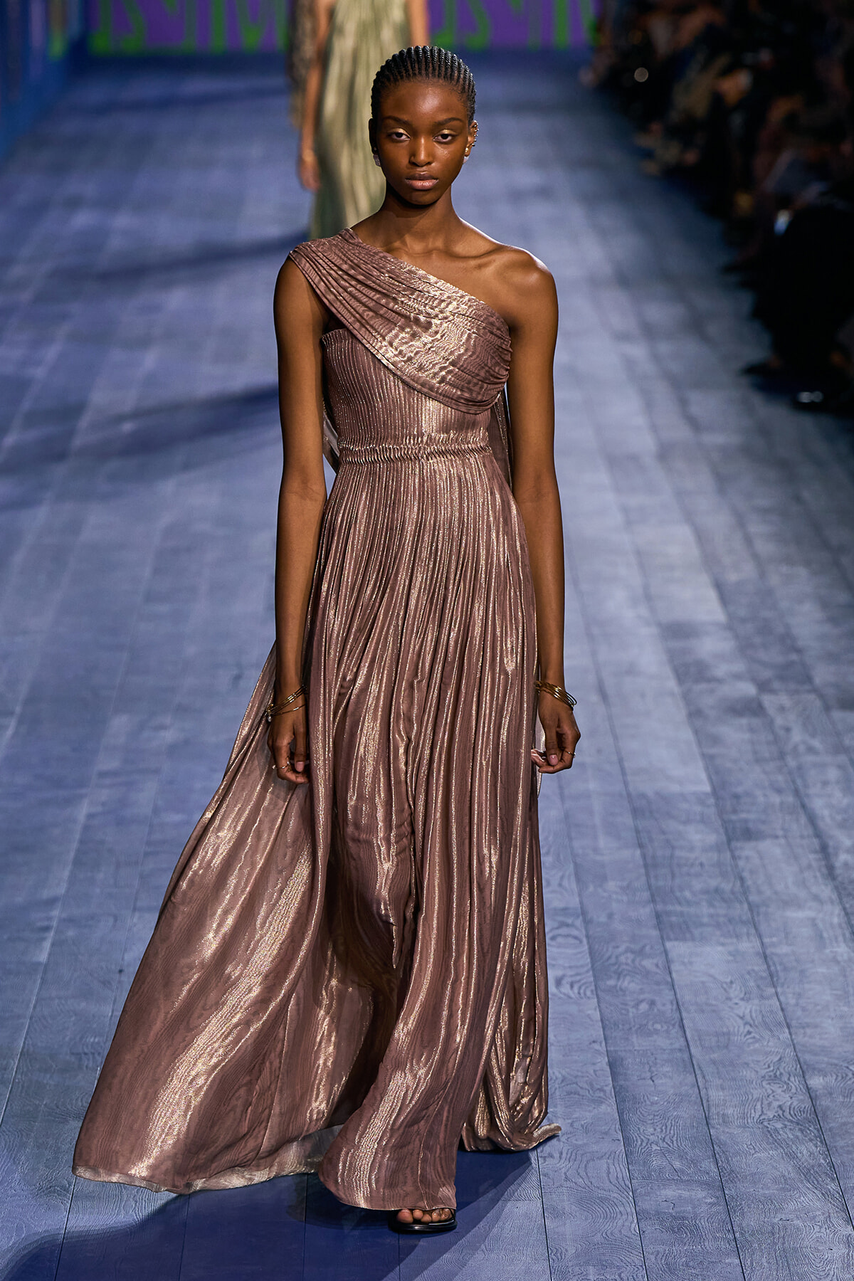 Model walking the runway in a metallic mauve one-shoulder gown with draped bodice and flowing pleated skirt on a blue-tinted catwalk.