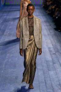 Model on a runway wearing a gold metallic blazer over a sheer brown top and bronze wrap-skirt with black sandals.