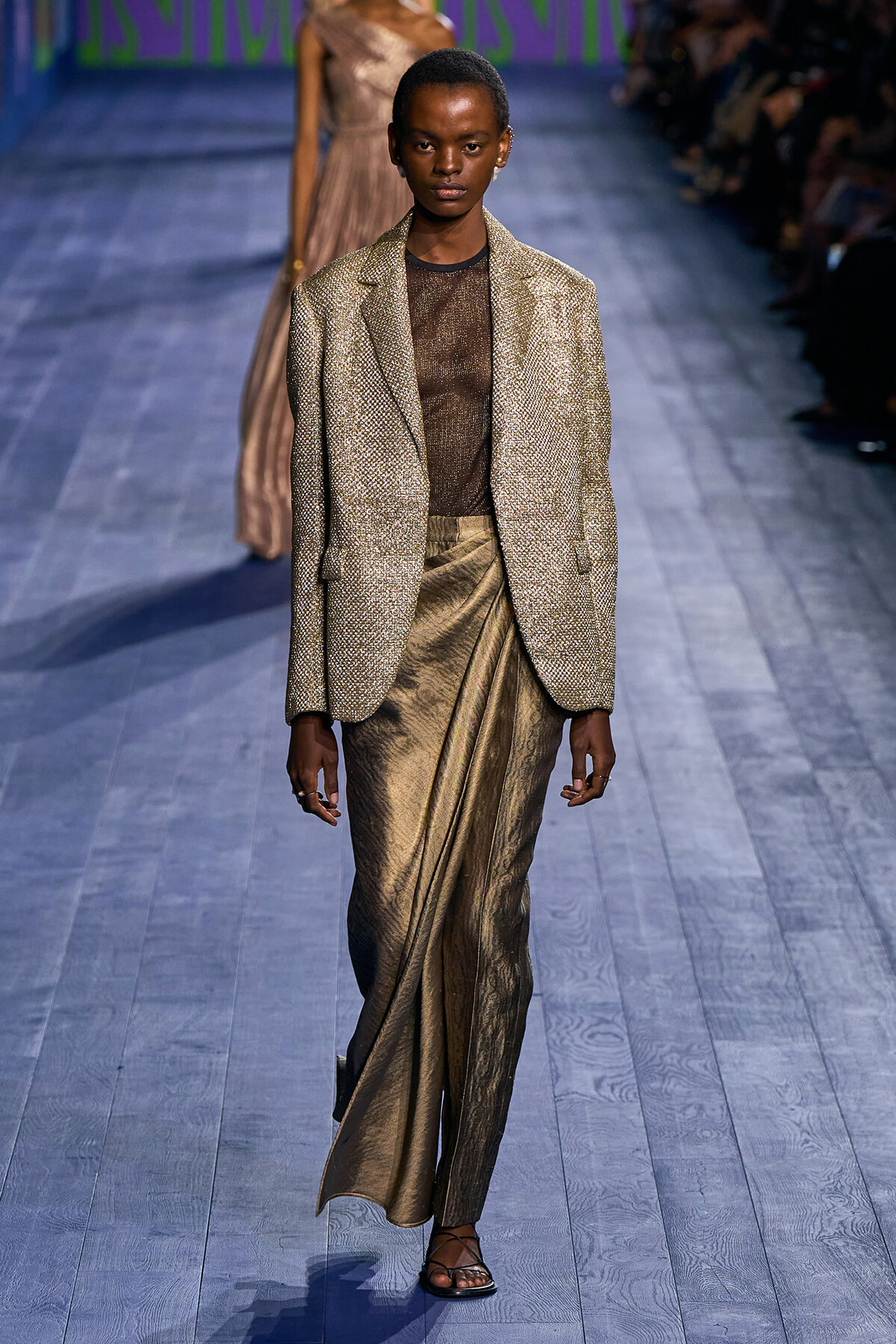 Model on a runway wearing a gold metallic blazer over a sheer brown top and bronze wrap-skirt with black sandals.