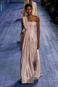 Runway model wearing a pale champagne one-shoulder, shimmery gown with draped bodice and floor-length skirt.