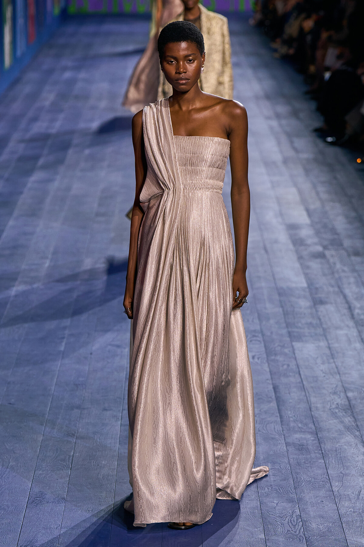 Runway model wearing a pale champagne one-shoulder, shimmery gown with draped bodice and floor-length skirt.