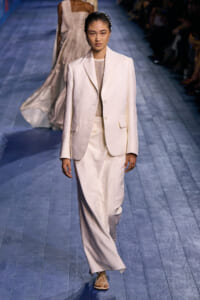Female model walks the runway in an off-white suit: tailored blazer and wide-leg trousers with a light top, hair slicked back.