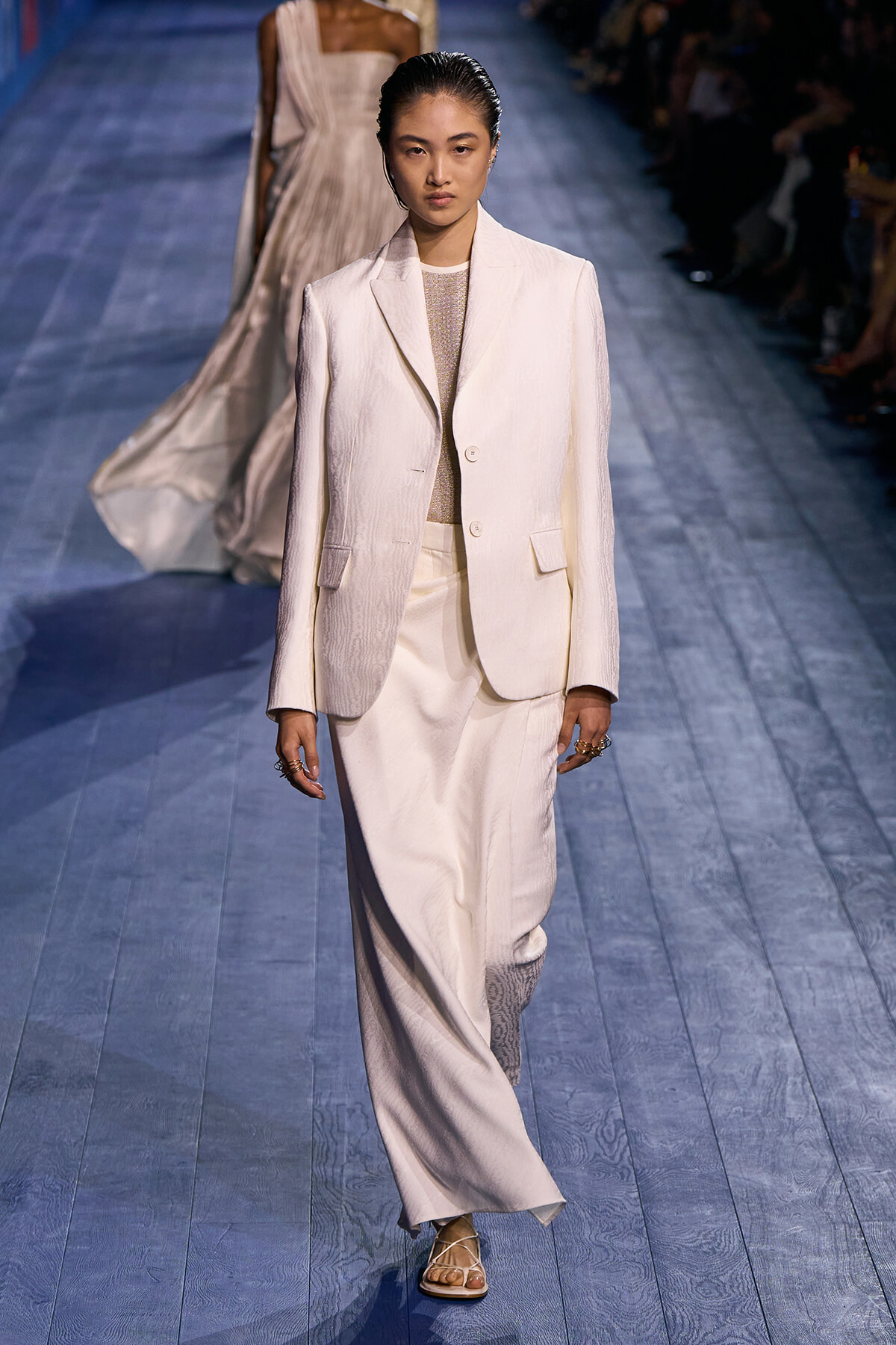 Female model walks the runway in an off-white suit: tailored blazer and wide-leg trousers with a light top, hair slicked back.