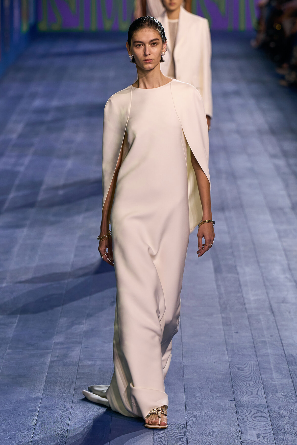 Model walking a runway in an ivory, cape-style gown with dramatic sleeve drape on a blue wooden catwalk, wearing sandals and bracelets.