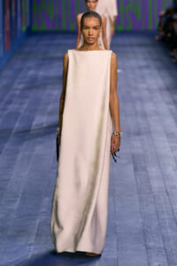Model walking a runway in a sleeveless ivory maxi dress with a high boat neckline and minimal jewelry.