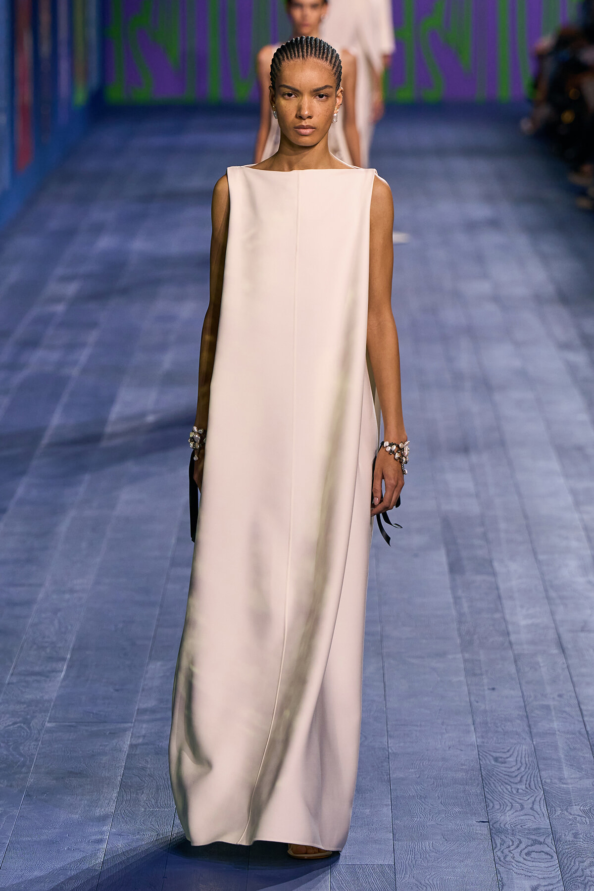 Model walking a runway in a sleeveless ivory maxi dress with a high boat neckline and minimal jewelry.