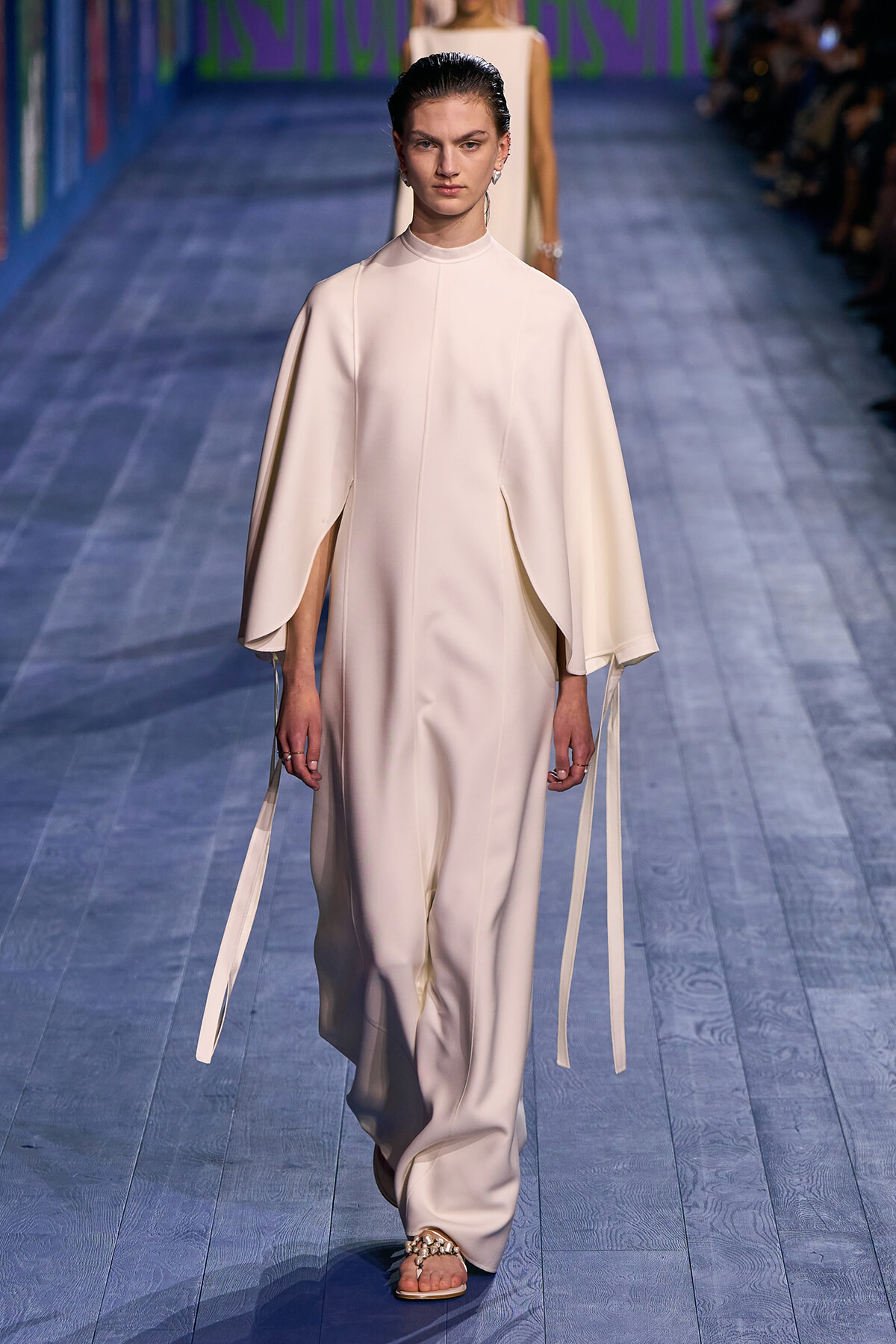 Model walks the runway in an ivory high-neck cape dress with dramatic flowing cape sleeves and long ribbon ties, paired with embellished flat sandals.