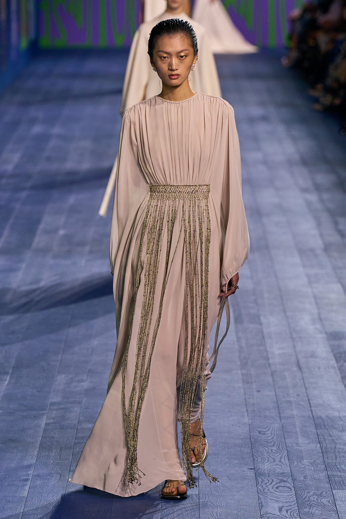 Model walks down a fashion runway in a pale pink gown with a gathered bodice and gold fringe at the waist.