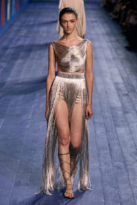 Runway model in a metallic gold fringe two-piece, with a long fringe skirt and strappy gladiator sandals.