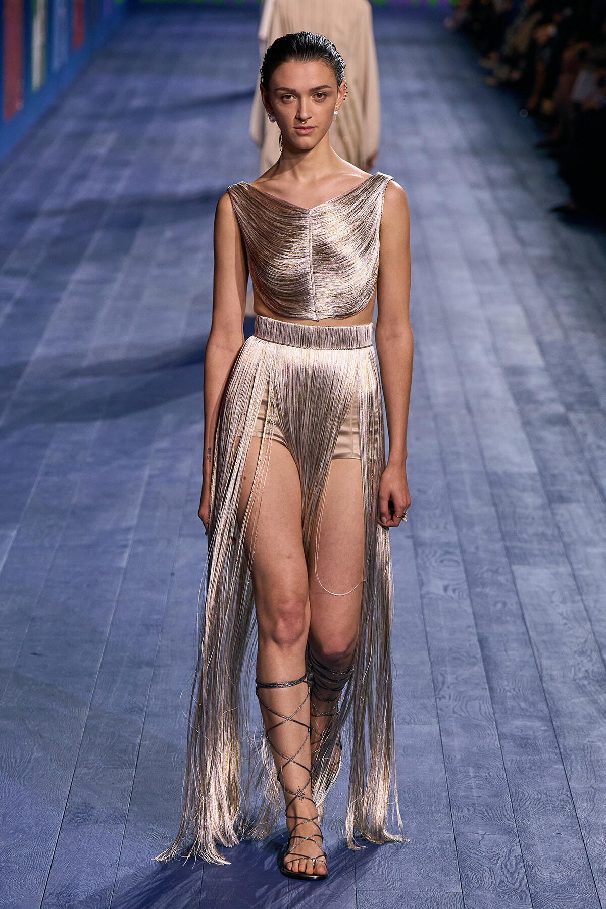 Runway model in a metallic gold fringe two-piece, with a long fringe skirt and strappy gladiator sandals.