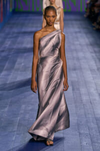 Female model walking a fashion runway in a metallic, one-shoulder satin gown with draped wrap detail on a blue wooden floor.