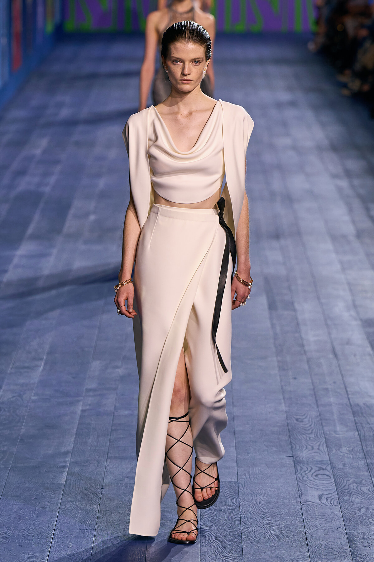 Model walking a runway in a cream draped crop top and wide-leg pants with a black sash belt and lace-up sandals.