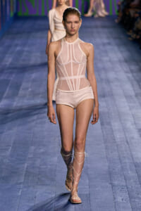 Model walks the runway in a pale pink sheer bodysuit with structured paneling and high neckline, paired with pale shorts and lace-up gladiator sandals.