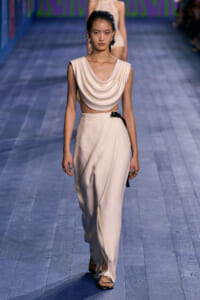 Model walks a fashion show wearing a cream draped sleeveless top and high-waisted wide-leg pants, with a black belt and strappy black sandals.
