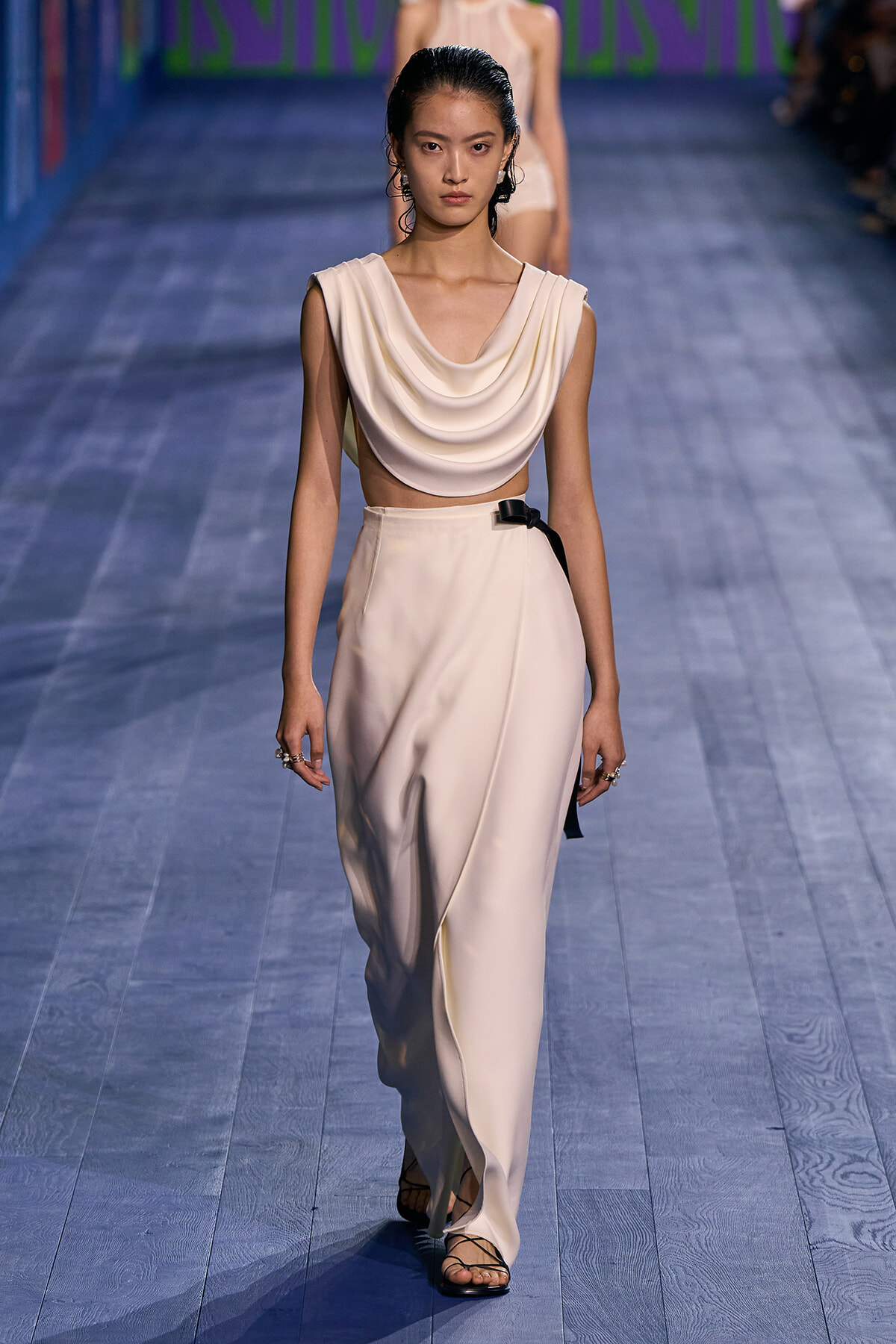 Model walks a fashion show wearing a cream draped sleeveless top and high-waisted wide-leg pants, with a black belt and strappy black sandals.