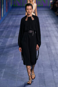Model walks a fashion runway in a black belted dress with cutout neckline and long sleeves, wearing lace-up sandals.