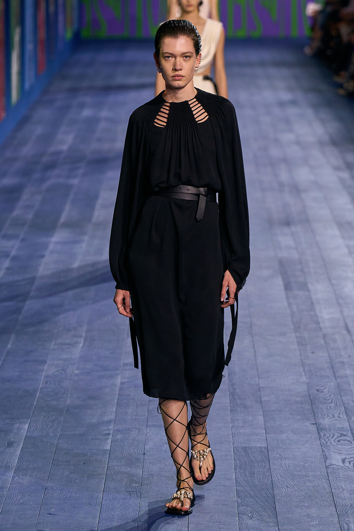 Model walks a fashion runway in a black belted dress with cutout neckline and long sleeves, wearing lace-up sandals.