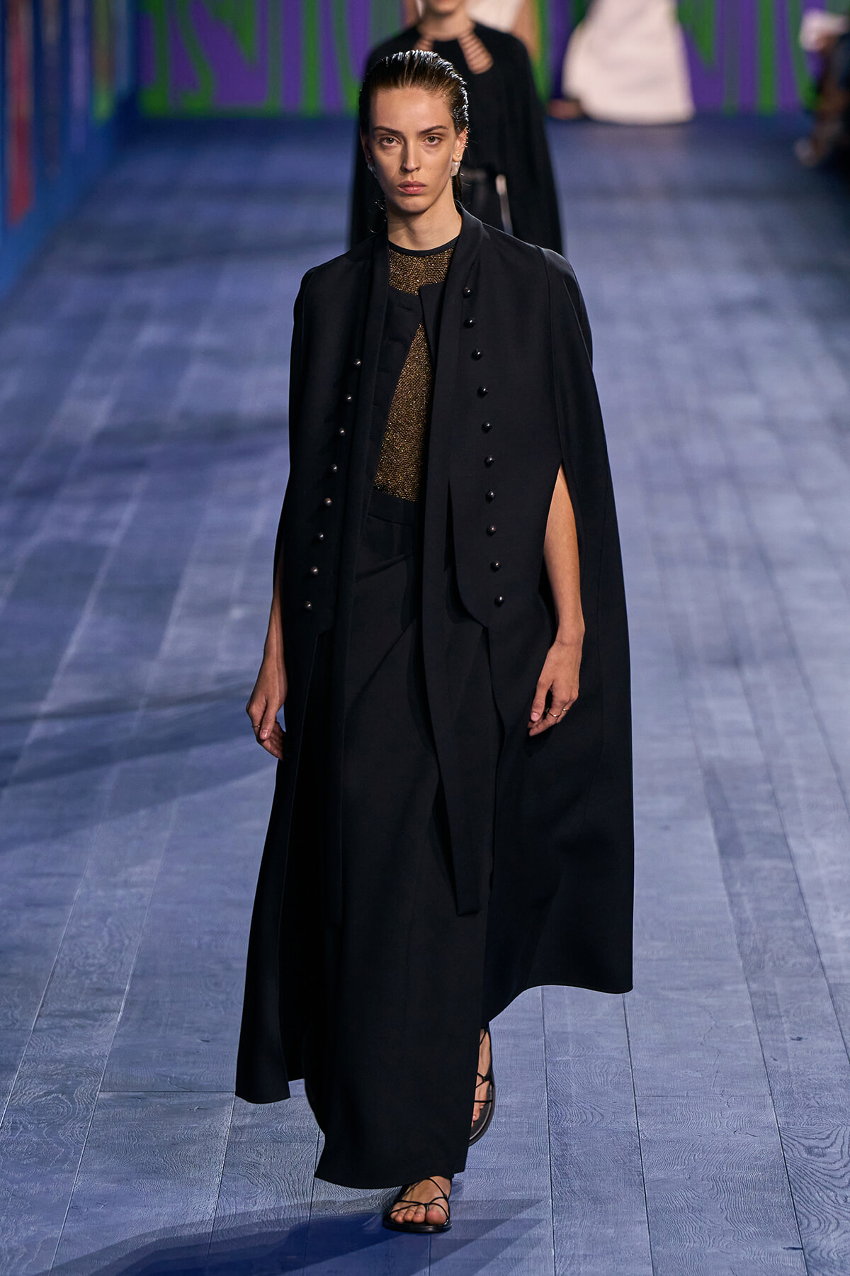 Model walks a runway wearing a long black cape over a tailored black suit with a metallic gold top underneath and flat sandals, on a blue-toned catwalk.
