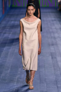 Female model walks a runway in an ivory sleeveless midi dress with a draped neckline and lace-up sandals.