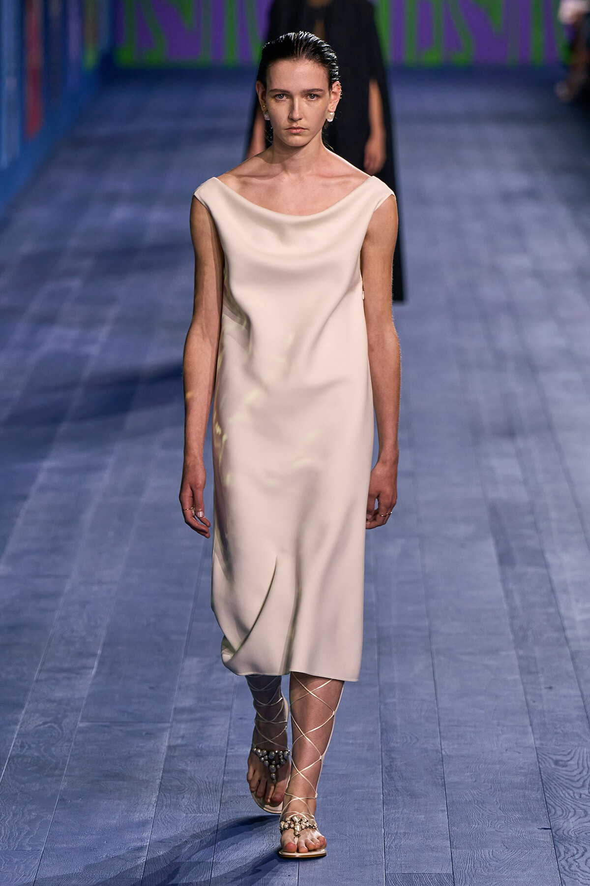 Female model walks a runway in an ivory sleeveless midi dress with a draped neckline and lace-up sandals.