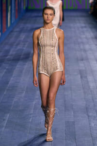 Model on a runway wearing a beige sleeveless perforated bodysuit with vertical stripes and lace-up sandals.