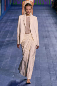 Model walks a cream tailored suit with a draped, pleated pants silhouette down a blue-toned runway.