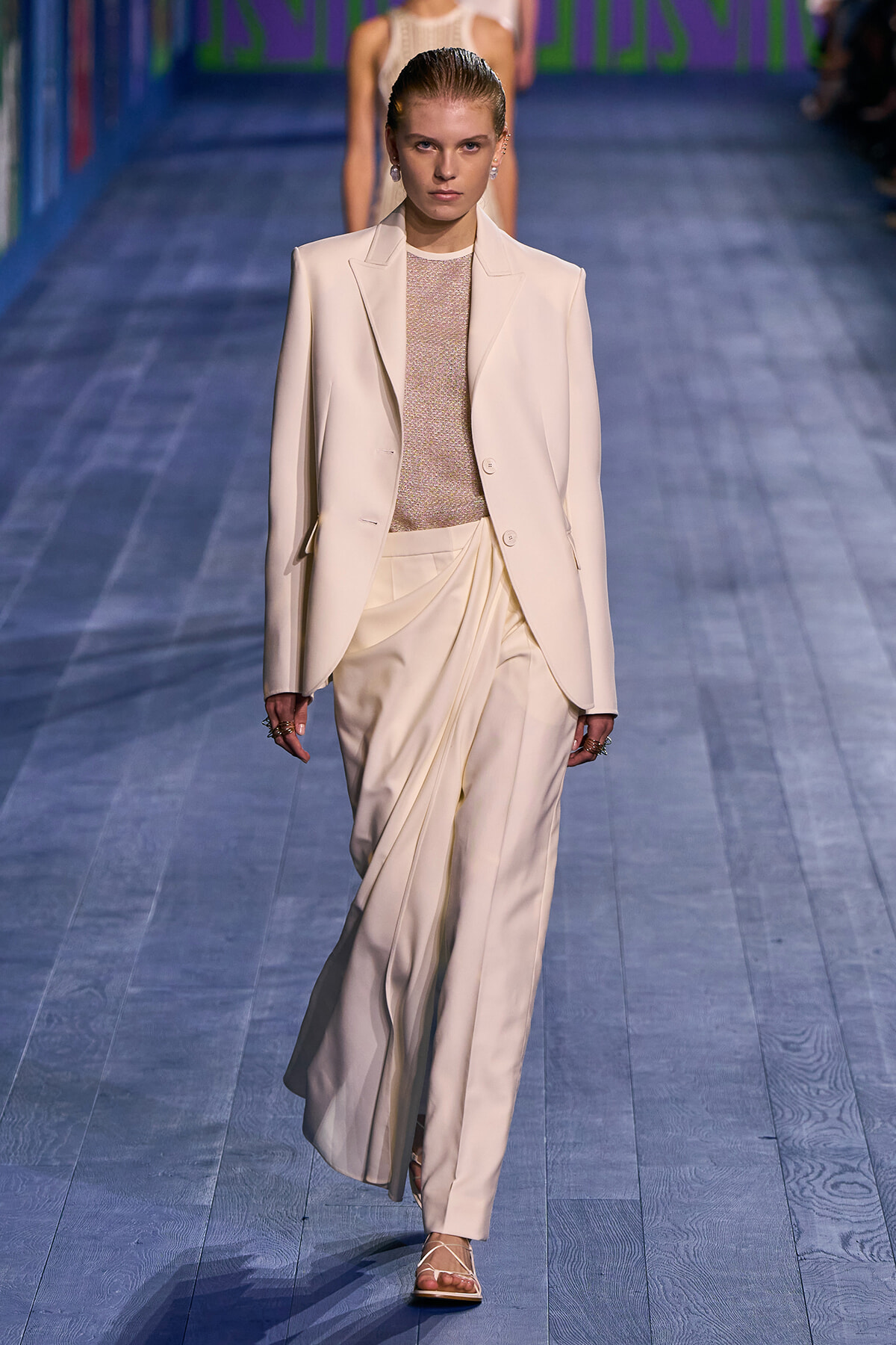 Model walks a cream tailored suit with a draped, pleated pants silhouette down a blue-toned runway.