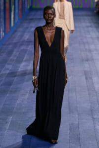 Model wearing a sleeveless deep-V black evening gown with a pleated fitted bodice and long flowing skirt, walking the runway with pearl bracelets.