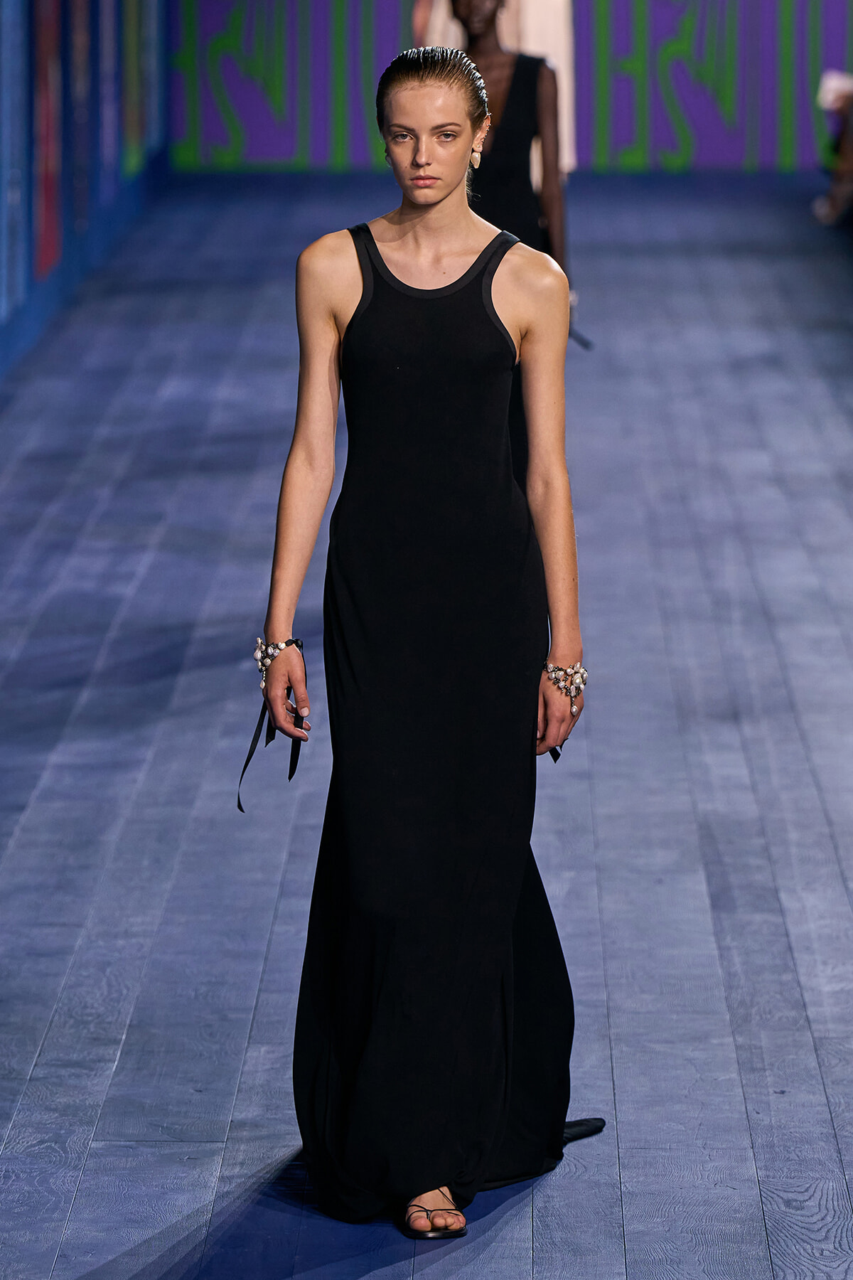 Model walks the runway in a black sleeveless maxi dress, hair slicked back, wearing bracelets and flat sandals on a blue-toned catwalk.
