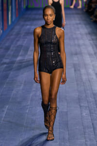 Model walks a fashion show wearing a black sleeveless sequined bodysuit with vertical stripe pattern and lace-up knee-high sandals on a blue runway.