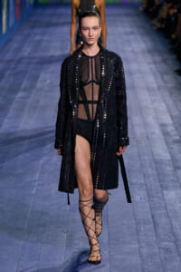 Model walking a fashion runway in a sheer black mesh bodysuit under a long sequined black coat with vertical embellishments and crisscross gladiator sandals