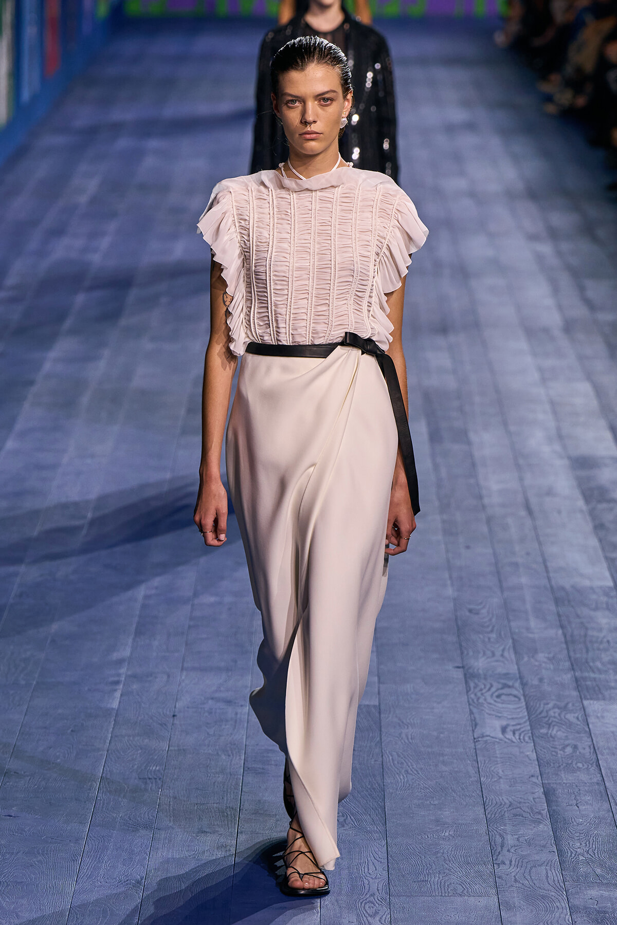 Model walking a runway in a pale pink ruched blouse with ruffled sleeves and cream wide-leg pants tied with a black belt on a blue wooden floor